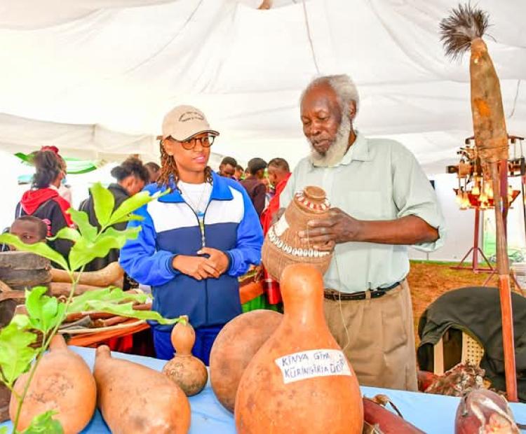 County Executive Committee Member for Trade, Cooperatives, Culture and Tourism, Diana Kendi (left) is taken through a demonstration of the names and functions of the Gikuyu tools by Mathenge Iregi